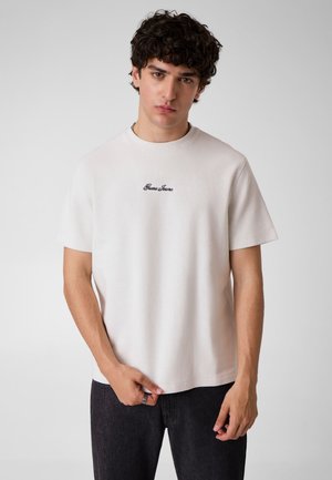 Jeune homme aux cheveux foncés et bouclés porte un t-shirt blanc uni Guess Jeans et un pantalon foncé, debout devant un fond gris clair.