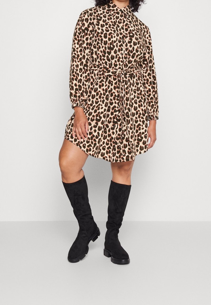 Femme portant une robe à manches longues imprimée léopard beige et noire avec une ceinture à la taille, associée à des bottes noires montantes, debout sur un sol uni.