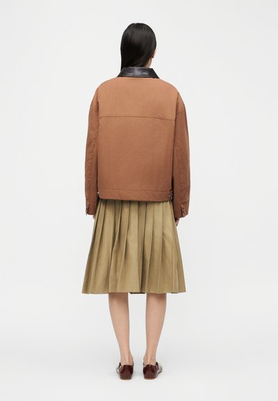 Γυναίκα με μακριά σκούρα μαλλιά που φορά καφέ μπουφάν με μαύρο γιακά, μπεζ πλισέ μίντι φούστα και καφέ παπούτσια, στέκεται με την πλάτη προς τα εμάς σε λευκό φόντο.