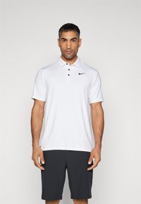 Hvid Nike polo-shirt med klassisk krave, tre sorte knapper og et lille sort logo. Kombineret med mørkegrå shorts.