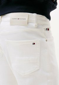 Jeans bianchi in denim con etichetta Tommy Hilfiger, caratterizzati da un piccolo logo e tasche standard. Texture liscia, design aderente.