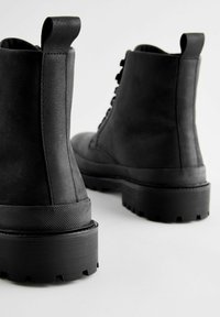 Botas de tobillo de cuero negro con un acabado texturizado, que cuentan con una suela de goma gruesa y una tira para facilitar ponerlas, exhibiendo cordones y un diseño de punta redonda.