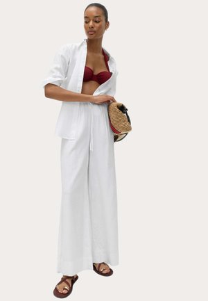 Kobieta ubrana w bordowy top bikini, oversize'ową białą koszulę, białe spodnie z szerokimi nogawkami, brązowe sandały, trzymająca plecioną torebkę.