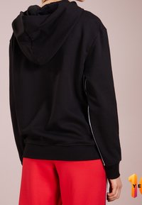 Černá mikina s kapucí a šňůrkami, s pruhy po stranách a volným střihem. Tkanina působí hladce, doplněna červenými tepláky.