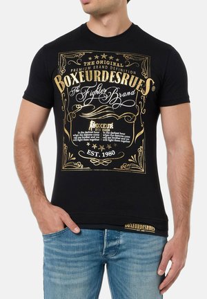 Schwarzes Baumwoll-T-Shirt mit goldfarbener Grafikdesign und Text sowie Schwirlen; kurze Ärmel und runder Ausschnitt, kombiniert mit hellen Jeans.