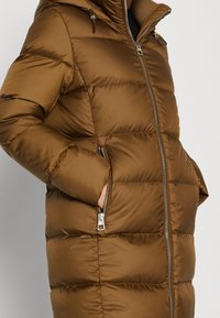 Bruine gewatteerde winterjas met een voorrits en met ritssluiting voorziene zijzakken, gedragen met de handen in de zakken, op een neutrale achtergrond.