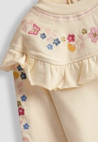 Cremfarbener Stoff mit rüschigen Kanten und floralem Stickerei in Rosa, Blau und Gelb, aus texturiertem Material.