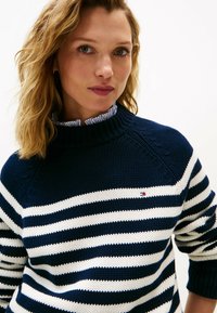 Frau mit schulterlangem blonden Haar, die einen navyblauen und weiß gestreiften Strickpullover über einem rüschengestalten Hemd trägt.