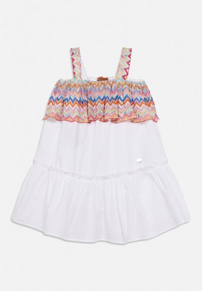 Λευκό φόρεμα χωρίς μανίκια με πολύχρωμο ζιγκ-ζαγκ μοτίβο στο ραμμένο μπούστο και τιράντες, με μια μικρή ασημί διακόσμηση κοντά στο κάτω μέρος.