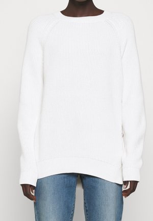 Osoba ubrana w biały sweter z prążkowanego dzianiny z długimi rękawami i niebieskie dżinsy, widoczna od ramion do górnej części ud.