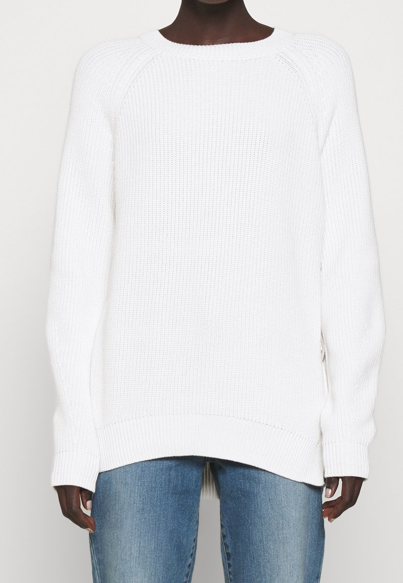 Personne portant un pull en maille côtelée blanche à manches longues et un jean bleu, visible des épaules jusqu'au haut des cuisses.