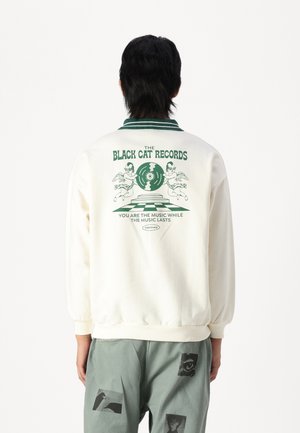 Sudadera color crema con estampado gráfico verde en la parte posterior, que presenta texto e ilustraciones. Cuello y dobladillo acanalados. Combinada con pantalones verdes.