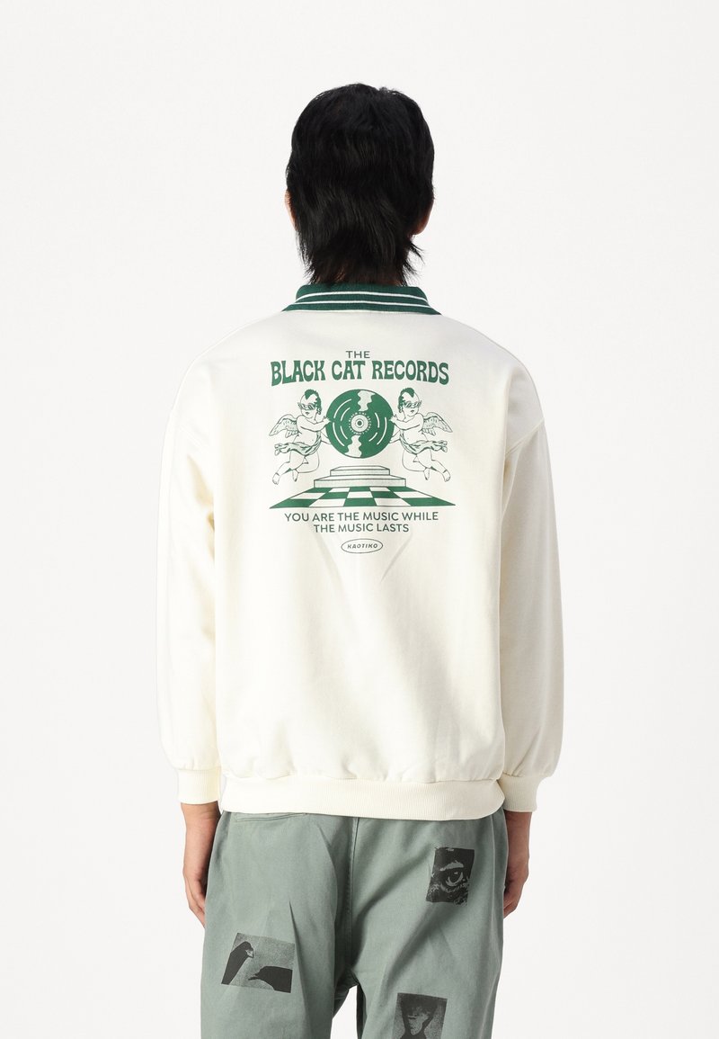 Kremowa bluzka z nadrukiem graficznym w kolorze zielonym na plecach, z tekstem i ilustracjami. Żebrowany kołnierz i dół. W zestawie z zielonymi spodniami.