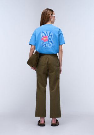 Frau mit braunen Haaren, die ein blaues T-Shirt mit Grafik, olivgrüne Hose, eine passende olivgrüne Tasche trägt und dunkle Schuhe anhat, von hinten gesehen.