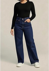 Personne portant un jean bleu foncé à jambes larges, un haut noir à manches longues, des baskets blanches, avec une main dans la poche, sur un fond uni.
