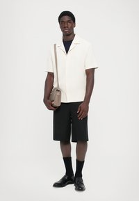 Herrenhemd mit kurzen Ärmeln in Off-White und einem Reverskragen, kombiniert mit schwarzen Shorts. Dazu gehören ein kleiner beiger Umhängetasche und schwarze Schuhe.