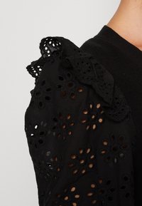 Svart blus med blommönster i hål, texturerad med rynkade axeldetaljer. Tyget verkar lätt och broderat.