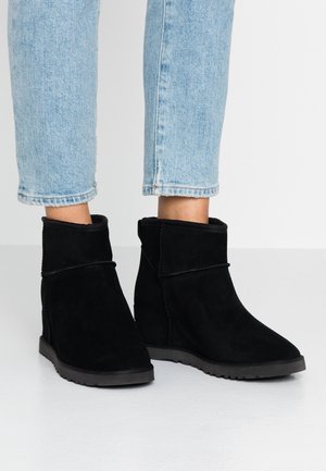 Person trägt hellblaue Jeans und schwarze Wildleder-Stiefeletten mit flachen Gummisohlen auf weißem Boden.