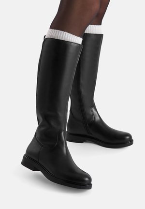 Botas negras de cuero hasta la rodilla con puntera redonda, cremallera lateral y tacón bajo. Presentan un puño de punto blanco en la parte superior. Textura suave.