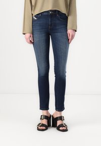 Jeans skinny de color azul oscuro con una textura suave y un ligero desvanecimiento, combinados con sandalias negras de tacón con acentos de hardware dorado.