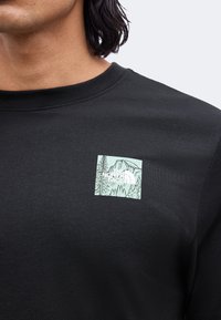 Κοντινή φωτογραφία ενός μαύρου T-shirt με μικρό τετράγωνο λογότυπο που δείχνει βουνά και δέντρα στο πάνω μέρος του στήθους κοντά στον γιακά.