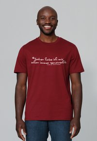 Ein lächelnder Mann trägt ein rotes Shirt mit dem deutschen Text "Sachen habe ich mir schon immer gewünscht" und steht vor einem schlichten grauen Hintergrund.