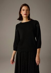 Pull en maille noire à manches trois-quarts, orné d'accents floraux en perles sur les épaules, associé à une jupe noire texturée.