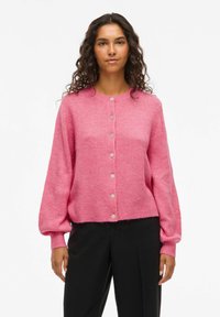 Femme aux cheveux bouclés portant un cardigan rose à boutons avec des manches bouffantes et un pantalon noir, se tenant devant un fond blanc uni.