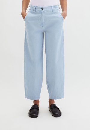 Personne portant un pantalon large bleu clair avec poches latérales et des mocassins noirs sans lacets, debout devant un fond uni.