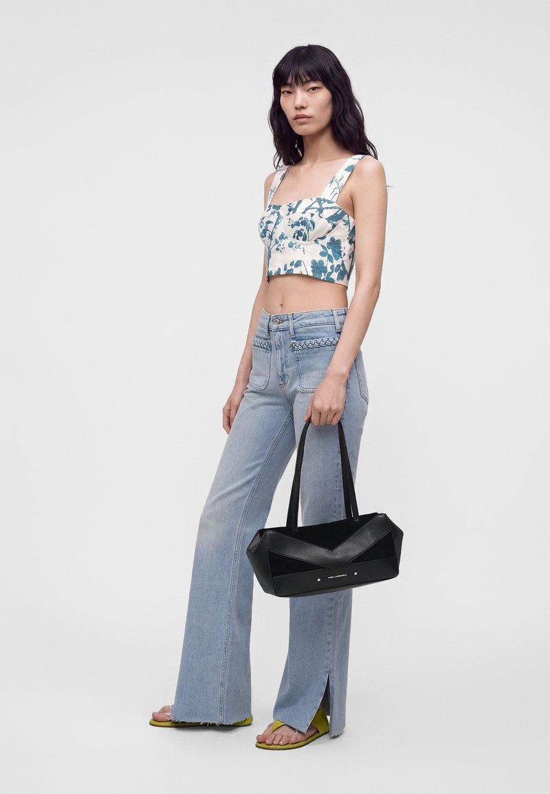 Femme portant un crop top fleuri et un jean évasé bleu clair, tenant un sac à main noir, debout sur un fond uni.