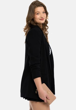 Black blazer with long sleeves, featuring a single button closure and pocket accents. Model holds a beige clutch purse. Simple urban background.