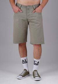 Khaki-Shorts mit geradem Schnitt, Vordertaschen, Metallknopf und kontrastierenden Nähten, kombiniert mit weißen Socken und grünen Turnschuhen.