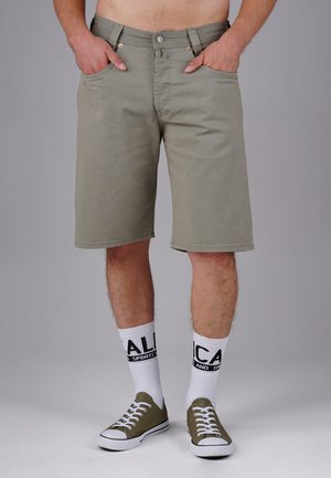 Khaki-Shorts mit geradem Schnitt, Vordertaschen, Metallknopf und kontrastierenden Nähten, kombiniert mit weißen Socken und grünen Turnschuhen.