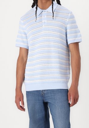 Homme portant un polo à manches courtes en maille à rayures bleu clair et blanc, associé à un jean en denim bleu moyen, debout devant un fond uni.