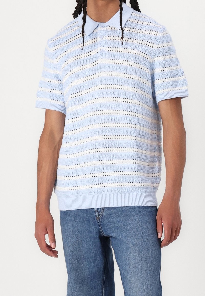Homme portant un polo à manches courtes en maille à rayures bleu clair et blanc, associé à un jean en denim bleu moyen, debout devant un fond uni.