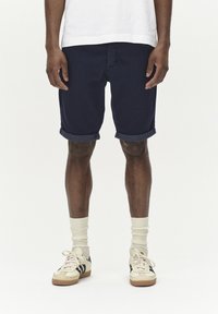 Pantaloni corti in cotone blu navy con risvolti, indossati con calzini crema e sneaker crema/nero. Design semplice, senza motivi visibili o accessori.