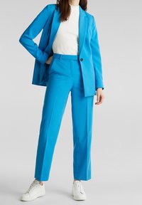 Modrý oblek s upraveným sakem a úzkými kalhotami, doplněný bílým strukturovaným topem a bílými teniskami. Hladká látka, elegantní design.