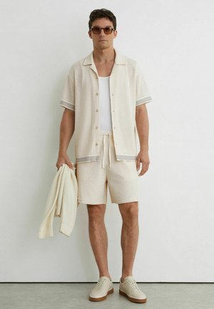 Bărbat purtând o cămașă bej cu mâneci scurte din plasă și pantaloni scurți, maiou alb, adidași bej, ținând un pulover asortat, stând lângă un perete alb.