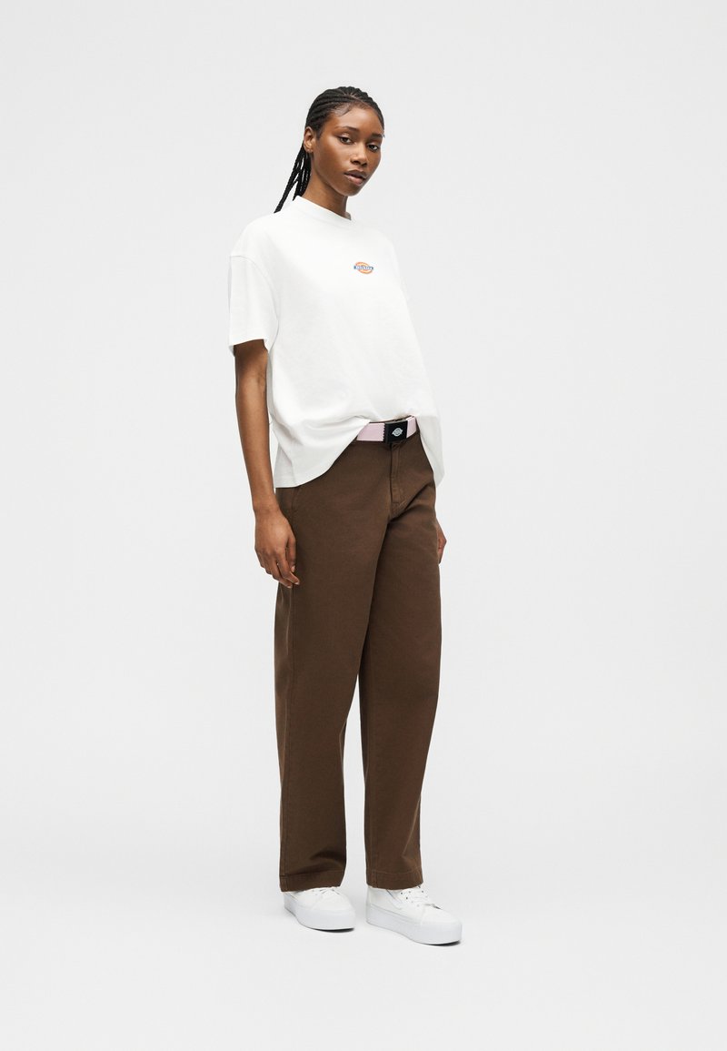 Donna con capelli intrecciati che indossa una maglietta bianca con logo, pantaloni marroni a gamba larga, scarpe da ginnastica bianche e una cintura rosa chiaro, in piedi davanti a uno sfondo semplice.