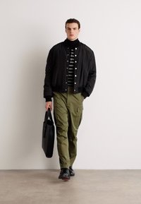 Schwarze Bomberjacke über einem gestreiften Rollkragenpullover, olivgrüne Cargo-Shorts und schwarze Schuhe. Trägt eine schwarze Tasche. Glatte Textur, lässiger Schnitt.
