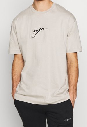 T-shirt beige à manches courtes avec logo "gpn" en cursive noire sur la poitrine, porté par une personne les mains dans les poches, associé à un pantalon noir.