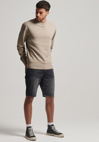 Felpa beige con scollo rotondo, abbinata a pantaloni corti in denim scuro e sneaker alte. La felpa presenta un discreto dettaglio del logo.