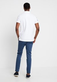 Vit polo med krage, korta ärmar och en logotyp på vänster axel; kombinerad med marinblå slim-fit byxor och marinblå skor.