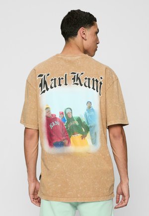 Tan oversized t-shirt featuring a large graphic print on the back of four individuals in colorful hoodies, with “Karl Kani” text in bold black.
