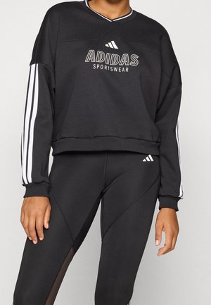 Person iført sort Adidas sweatshirt med hvide striber på ærmerne og matchende sorte leggings med logoer nær taljen.