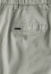 Světlé šedé kraťasy vyrobené z hladkého materiálu, s zadní kapsou a černou značenou nášivkou a viditelnými švy.