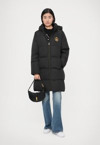 Melnā pūdera jaka ar kapuci, dimanta raksta dizains, zelta rāvējslēdzējs un emblemāts. Kombinēta ar izliektām džinsa biksēm un mazu melnu somiņu.