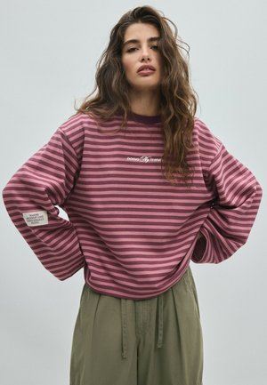 Frau mit langen welligen Haaren, trägt einen lila-rosa gestreiften Pullover mit Text, kombiniert mit lockeren olivgrünen Hosen, steht mit den Händen in der Hüfte.
