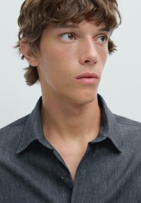 Junger Mann mit hellbraunen Haaren und braunen Augen, der ein dunkelgraues Hemd mit Kragen trägt und leicht nach rechts schaut, vor einem schlichten Hintergrund.