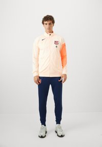 Men's track jacket in light peach with orange sleeve, featuring a logo on the chest. Paired with navy pants and gray athletic shoes.